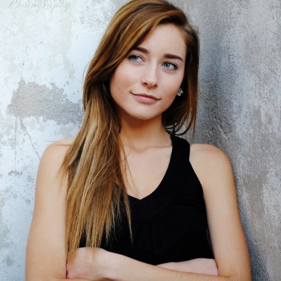 Woman with light brown hair leaning against gray wall, wearing blank tank.